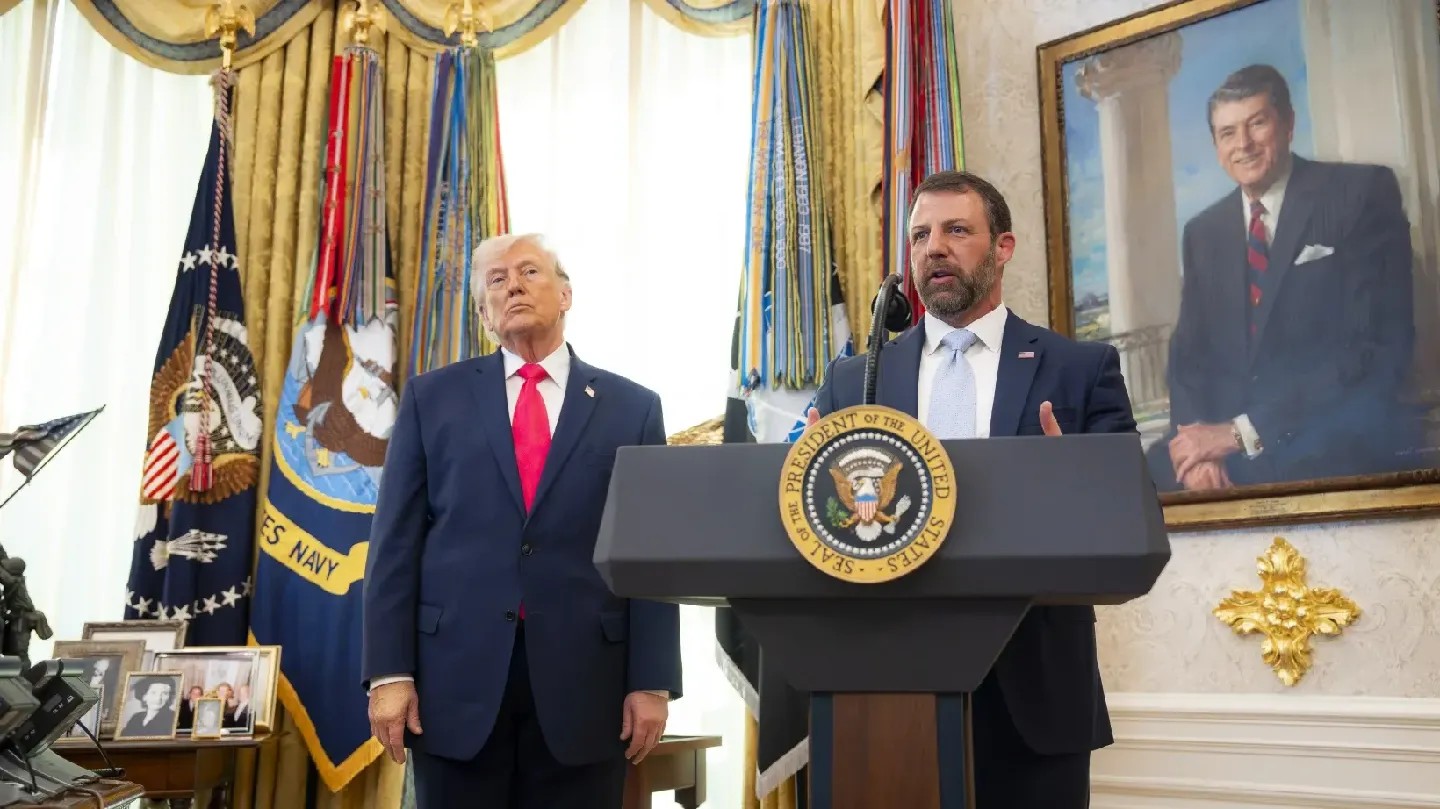 Mullen, at right, stands and speaks at a podium, while Trump, at left, looks on.