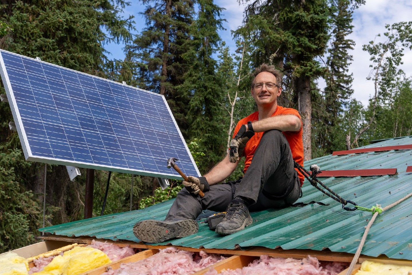 Blume-Kohout, holding a hammer, sits on a green roof, with pine trees in the background.