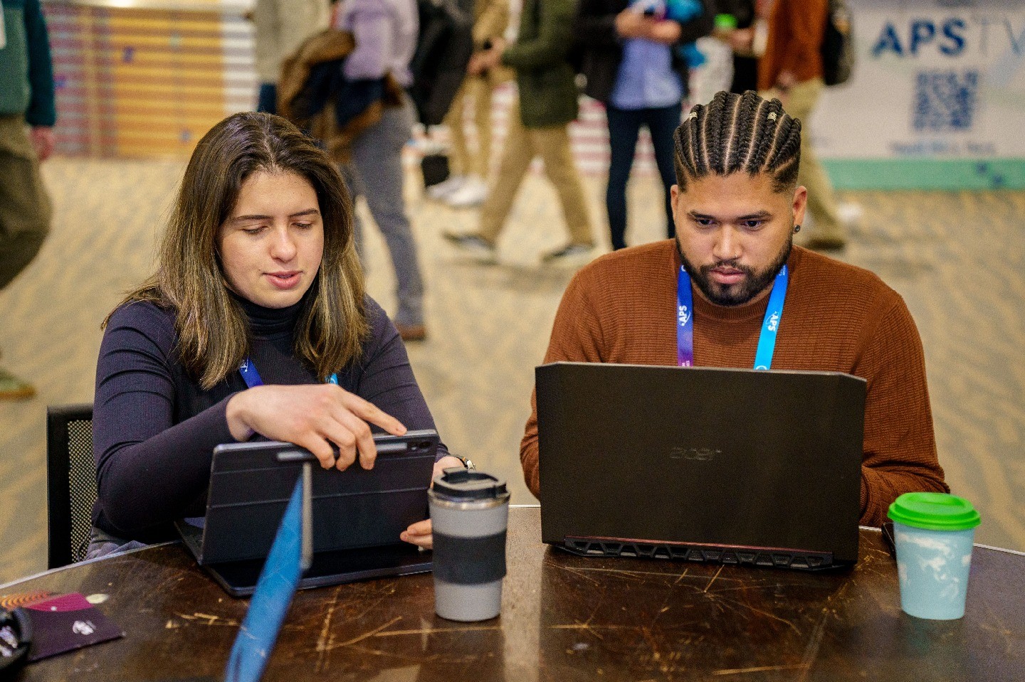 A woman and a man sitting at a table together and looking at a tablet and laptop