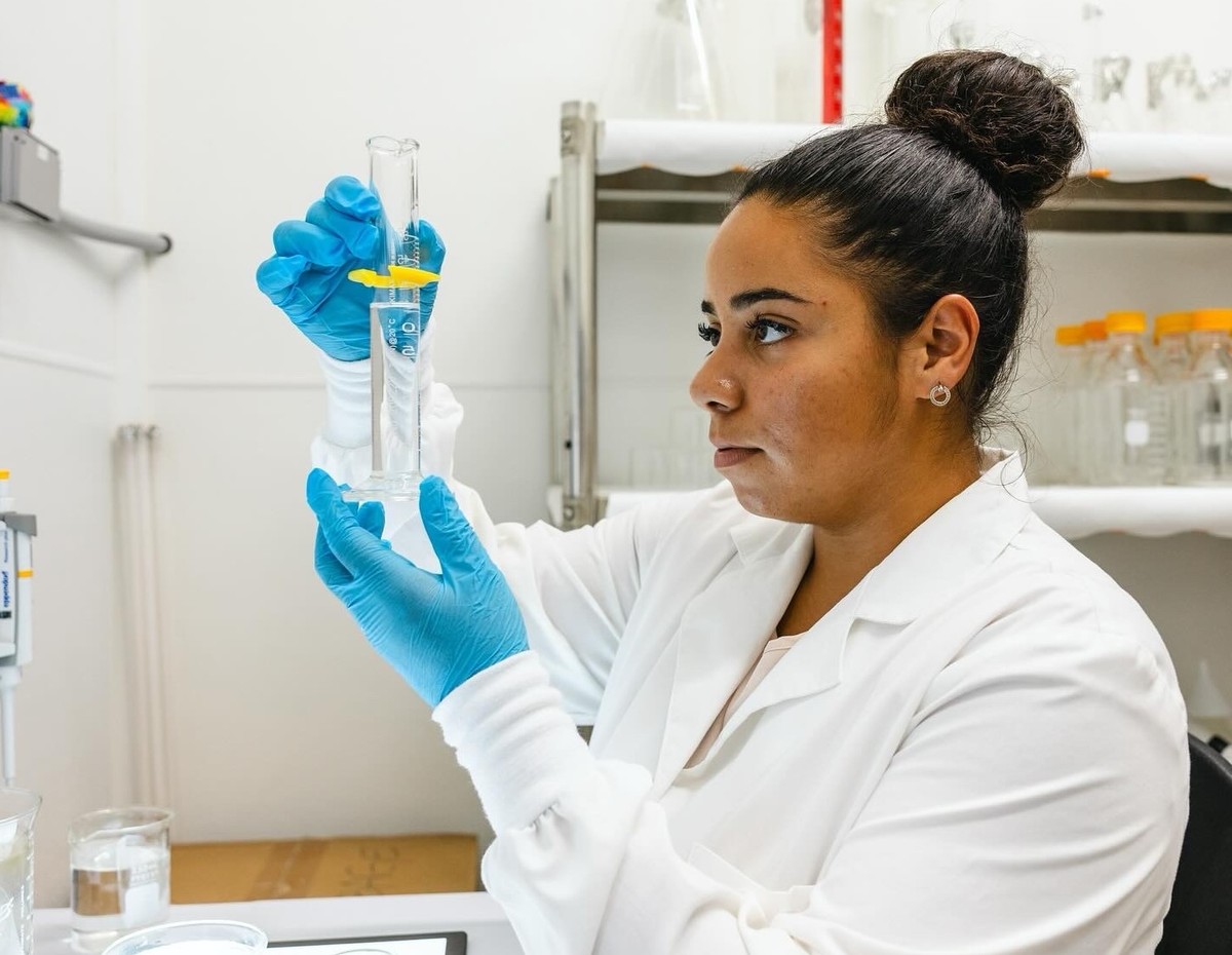 Rivera, wearing a white lab coat, holds up a beaker with liquid in it in a lab setting.