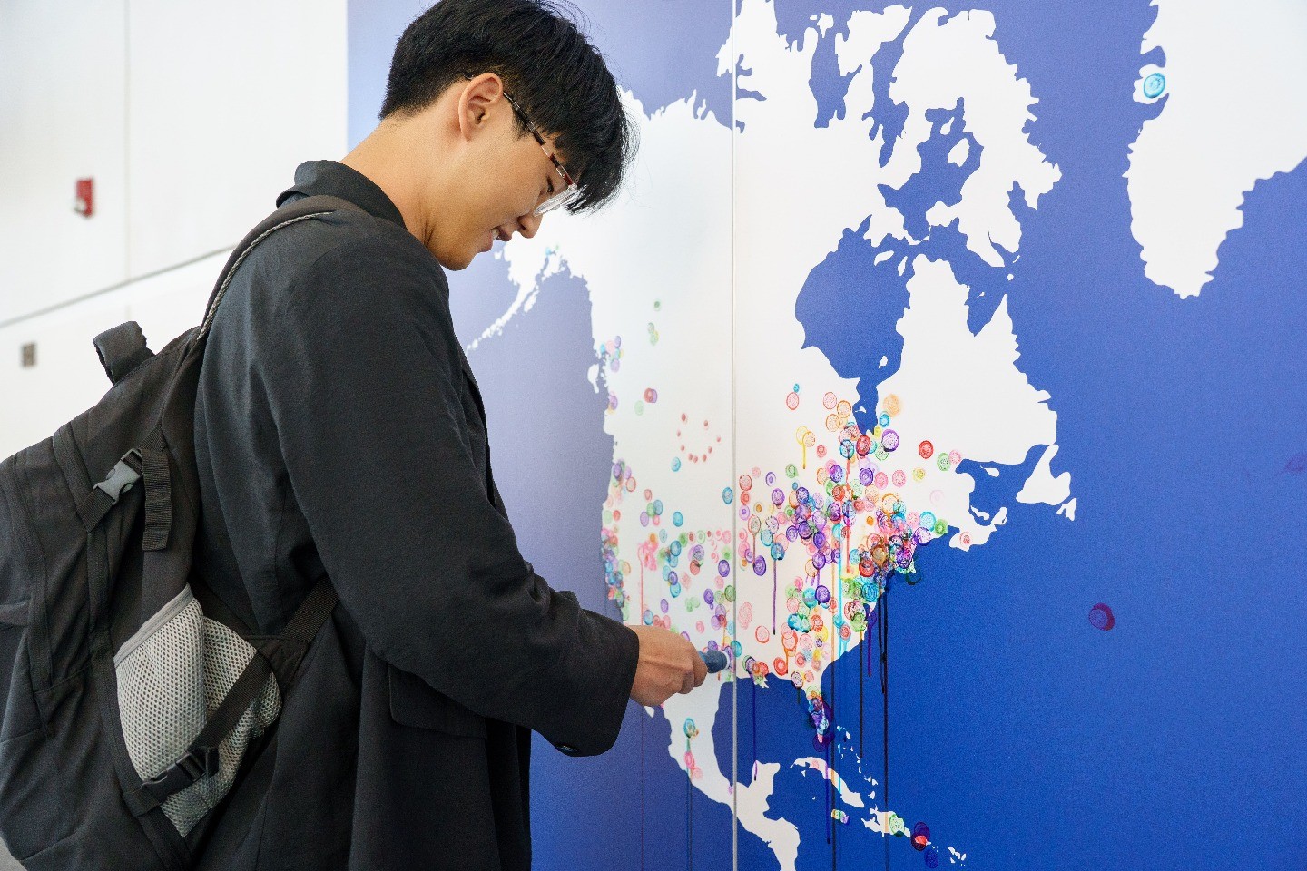 A person wearing a backpack stands and smiles in front of a map and places a pin on the map.