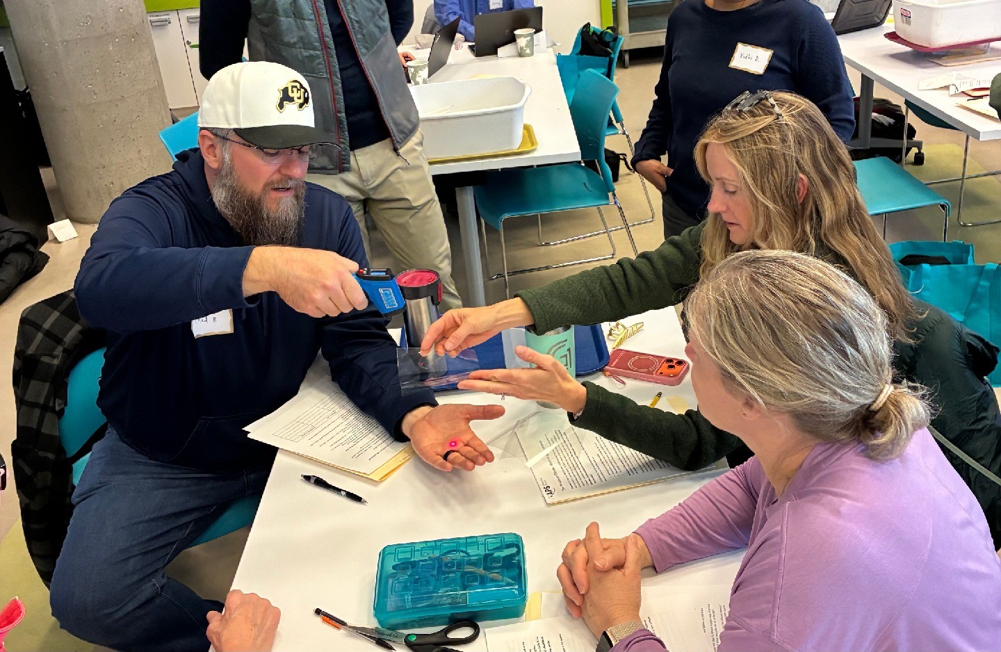 Three teachers sit at a desk; one teacher uses a device to measure the temperature of his palm while two other teachers look on.