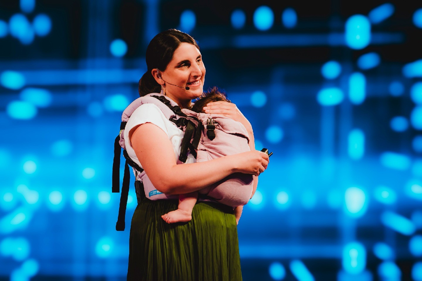 Carla Hermann Avigliano mic’ed up on stage with an infant in a carrier on her chest.