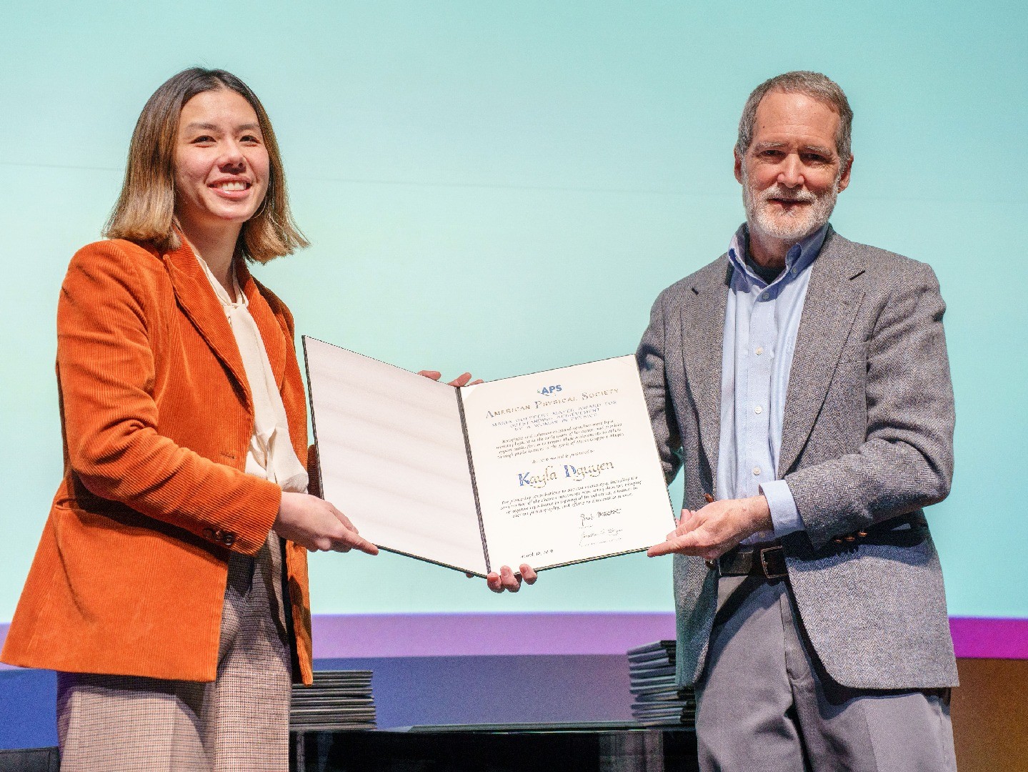 Two people on a stage each holding an award certificate open together as they pose for a photo.