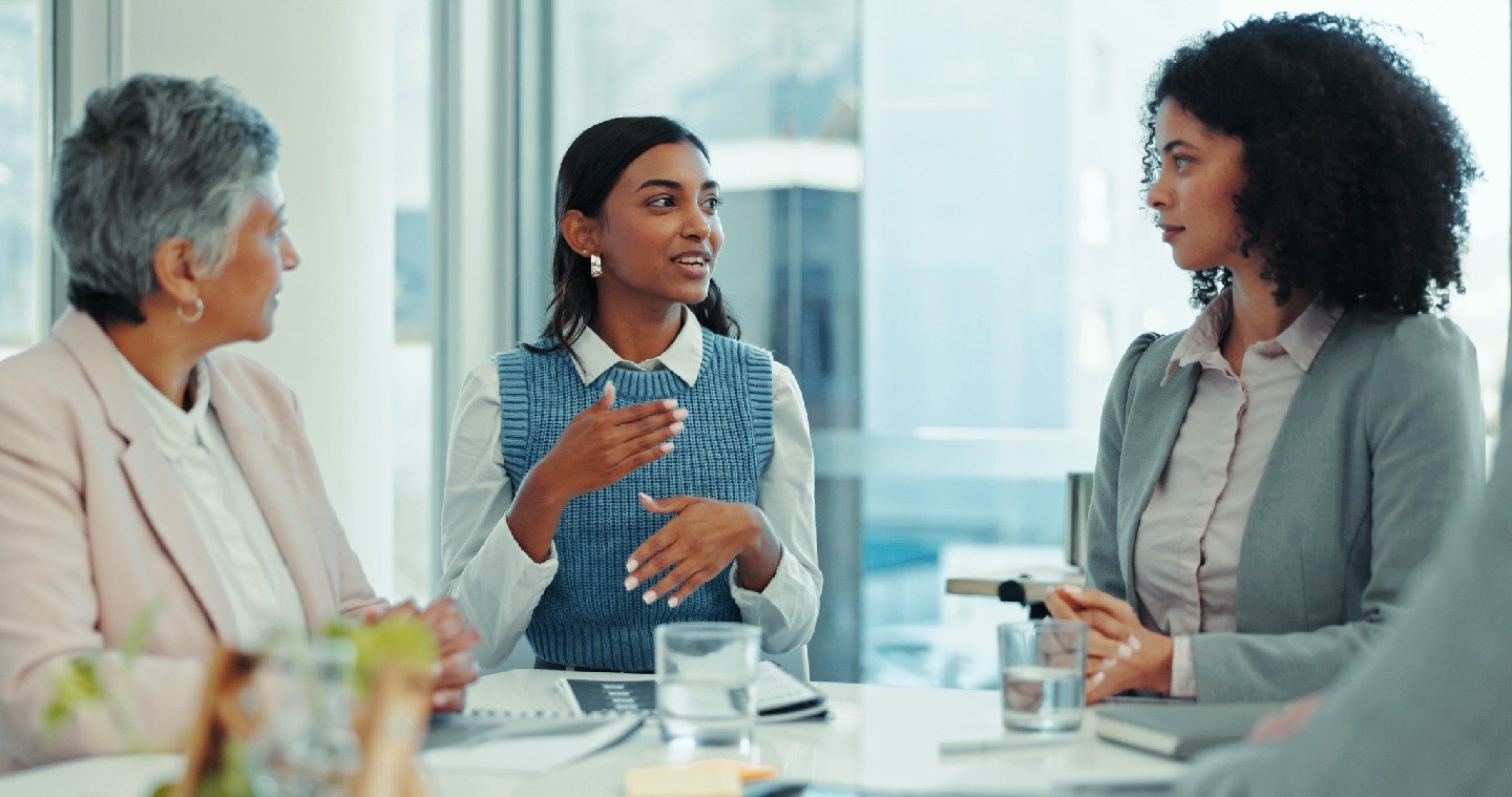 Three women in business attire seated at a table in conversation.