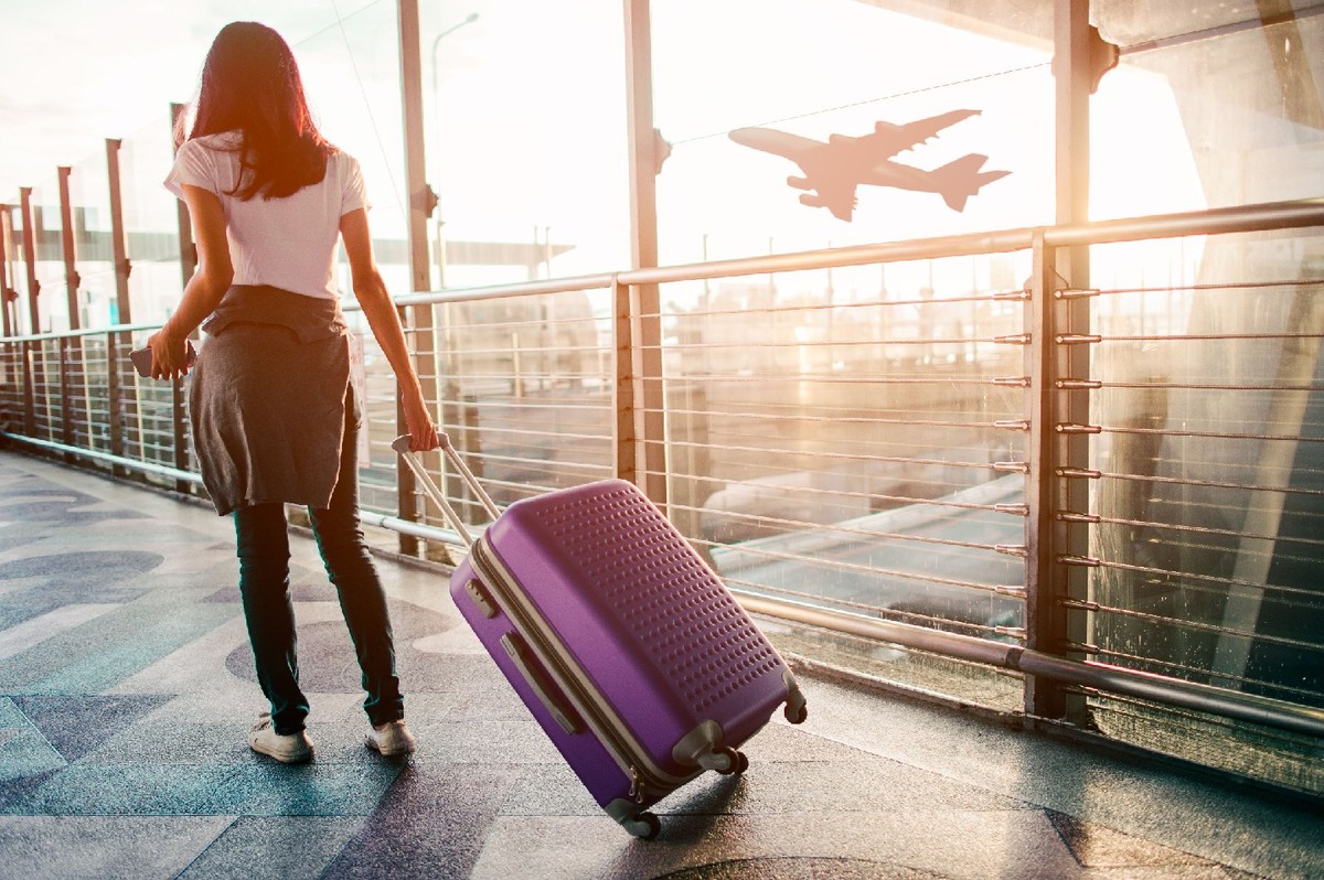 A person with rolling luggage inside an airport. A plane is in midair in the background.