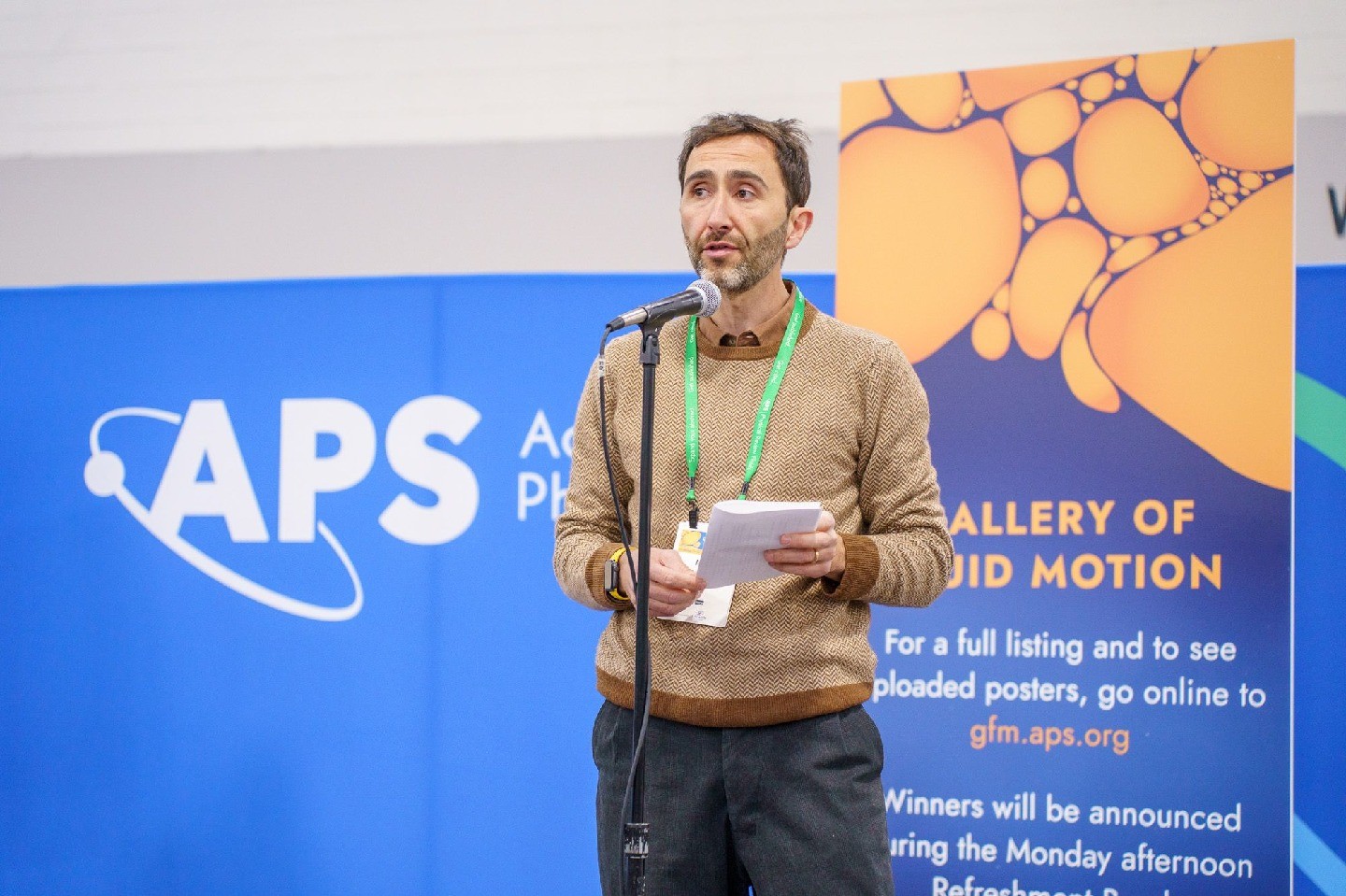 A person standing in front of a microphone. Behind them is an APS branded banner and a sign for Gallery of Fluid Motion