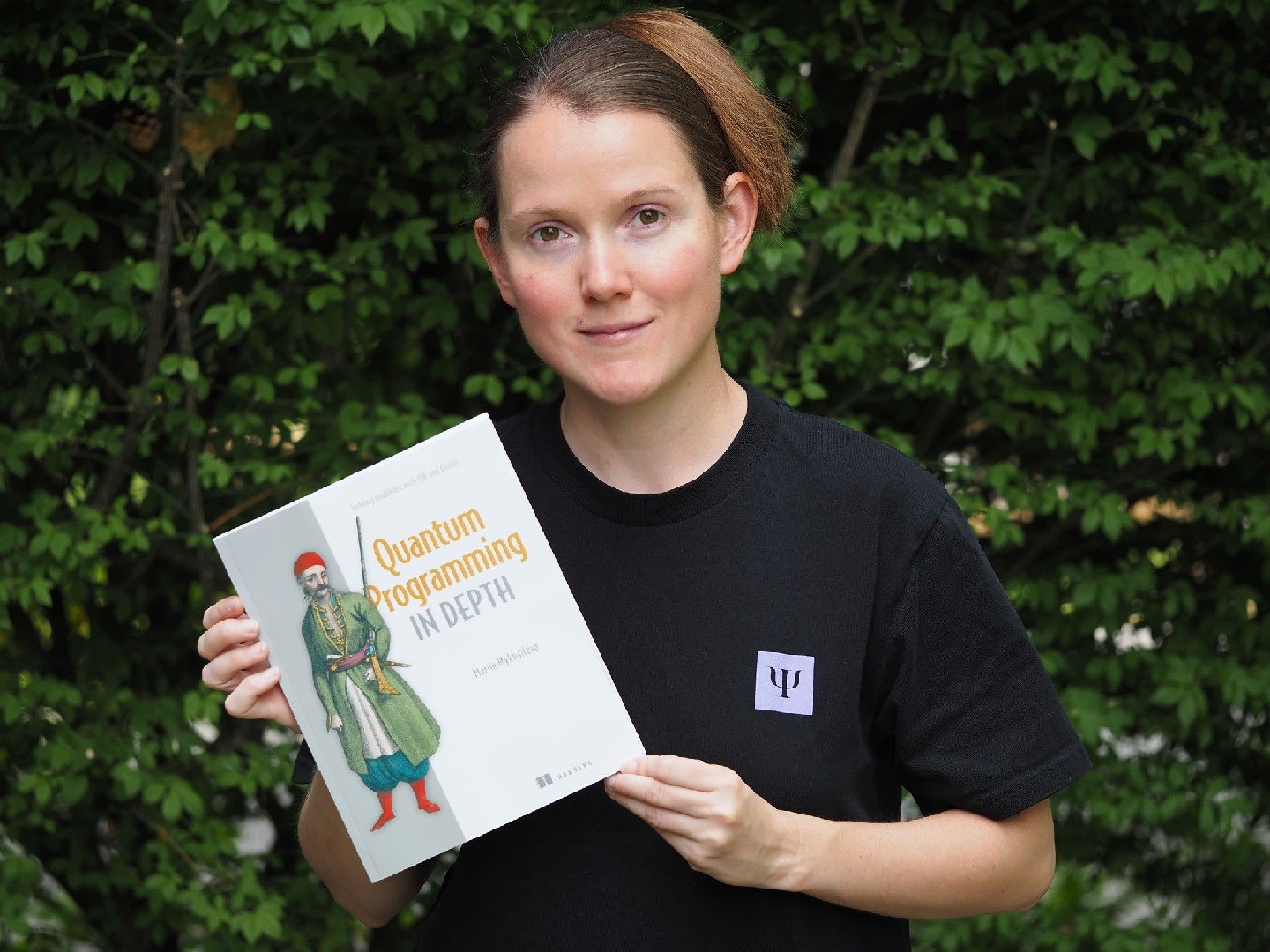 Mariia Mykhailova, wearing a black t-shirt and standing in front of green foliage, holds up her book, Quantum Programming in Depth.