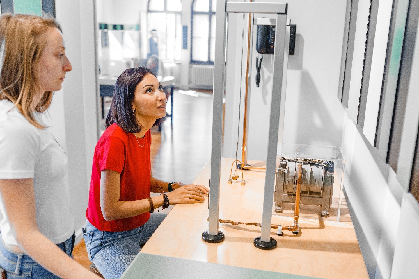 Two young women, one seated and one standing, study a physics experiment on the table in front of them.