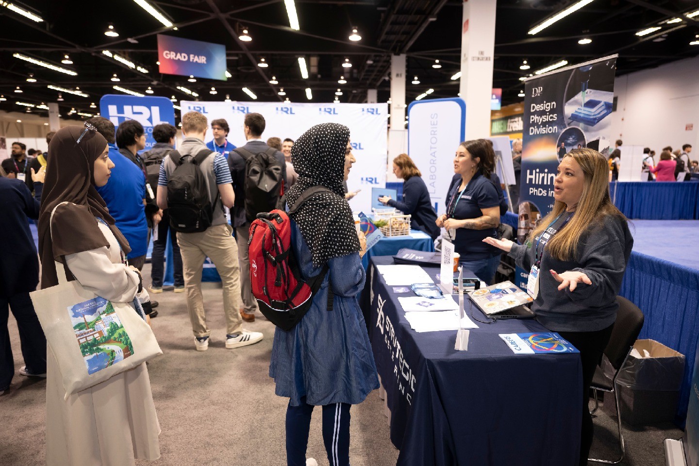 Exhibitors speaking to attendees at the APS Global Physics Summit exhibit hall, with a focus on two attendees wearing headscarves