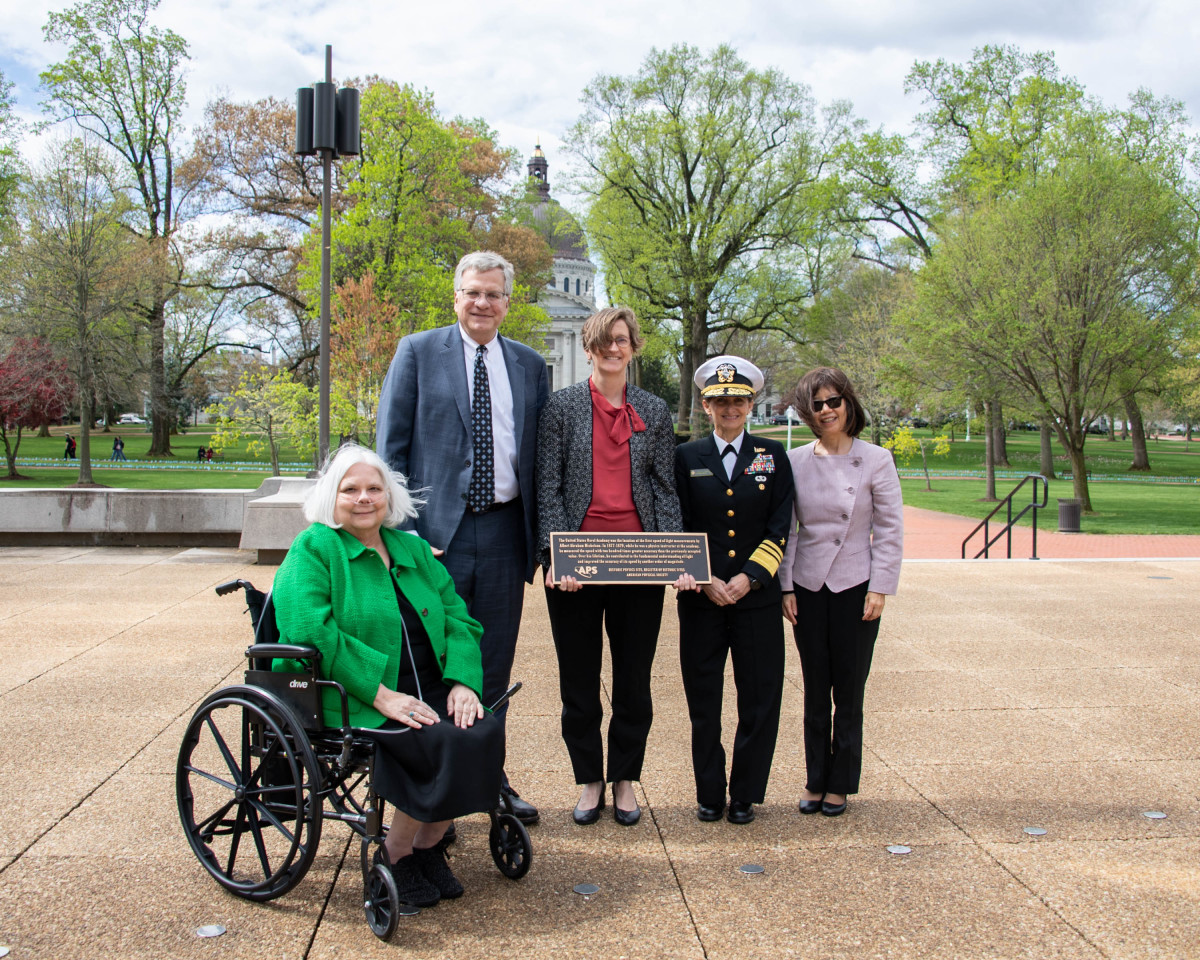 Five people -- including the APS President and CEO -- pose for a camera with a black-and-gold commemorative plaque signifying an APS Historic Site. They're standing outdoors on a plaza, with the U.S. Naval Academy campus visible behind them.