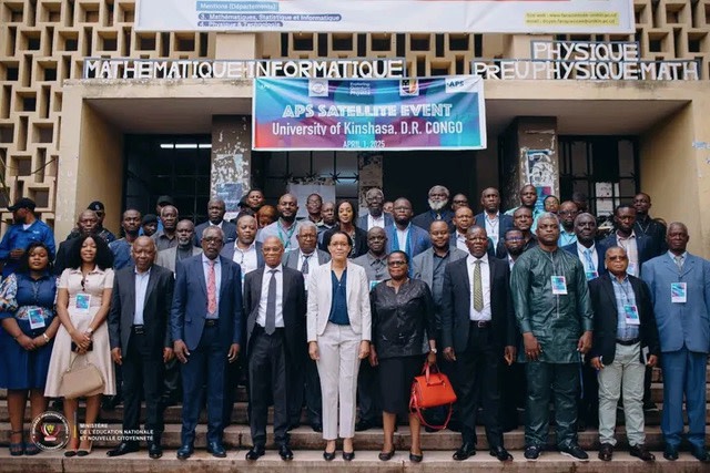 A crowd of meeting attendees at the satellite site in Congo pose in a group out front of a building, with an APS event sign hanging above them.