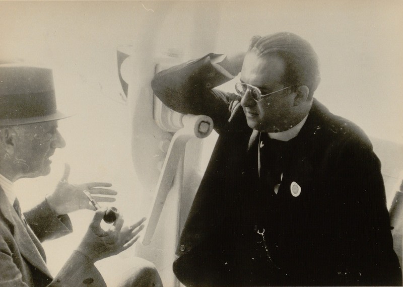 Arthur Eddington (at left), wearing a hat and holding a pipe, gesticulates in conversation with Georges Lemaître, who is wearing a dark suit and glasses.