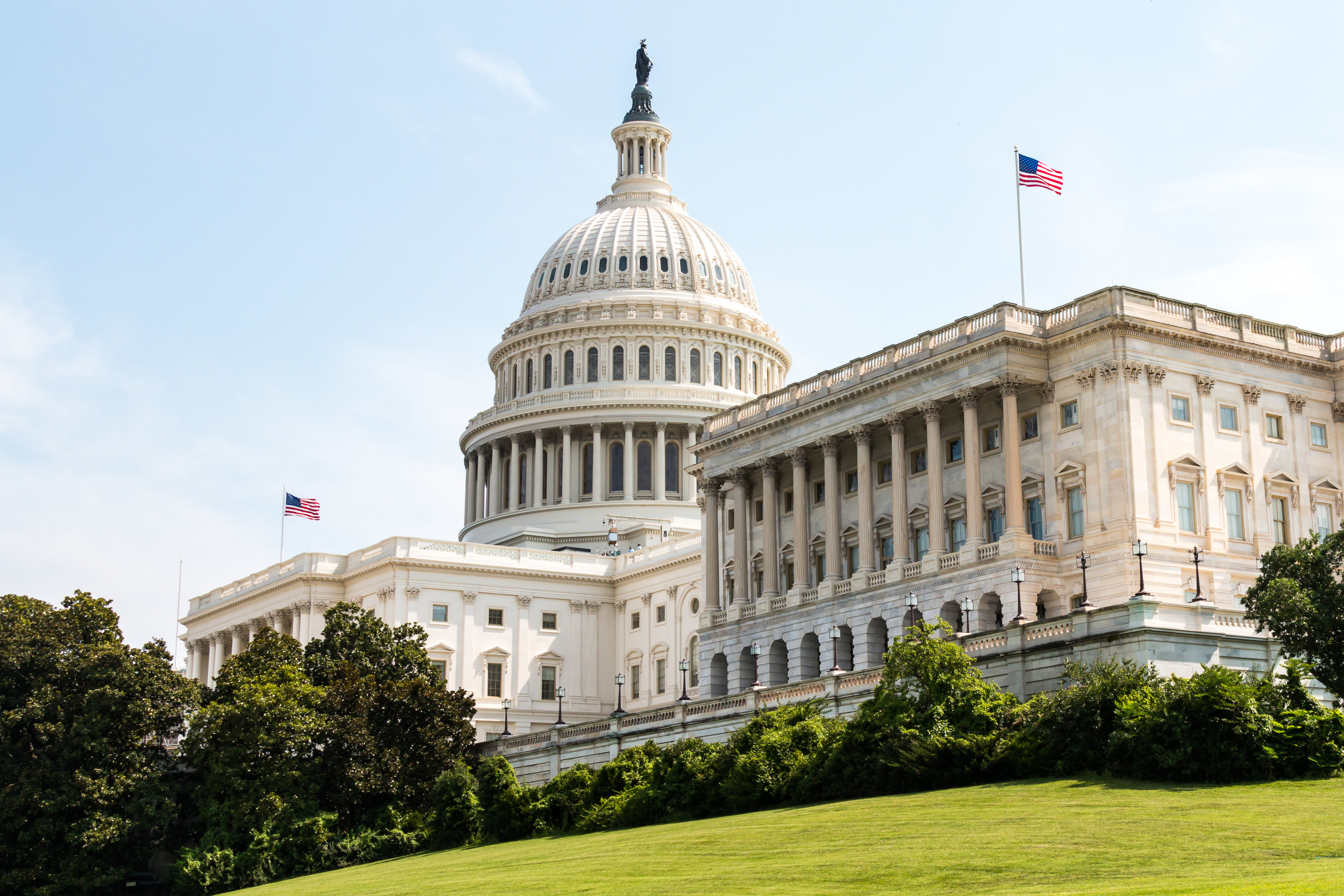 The U.S. Capitol building from a side angle, with the green lawn and blue sky visible.