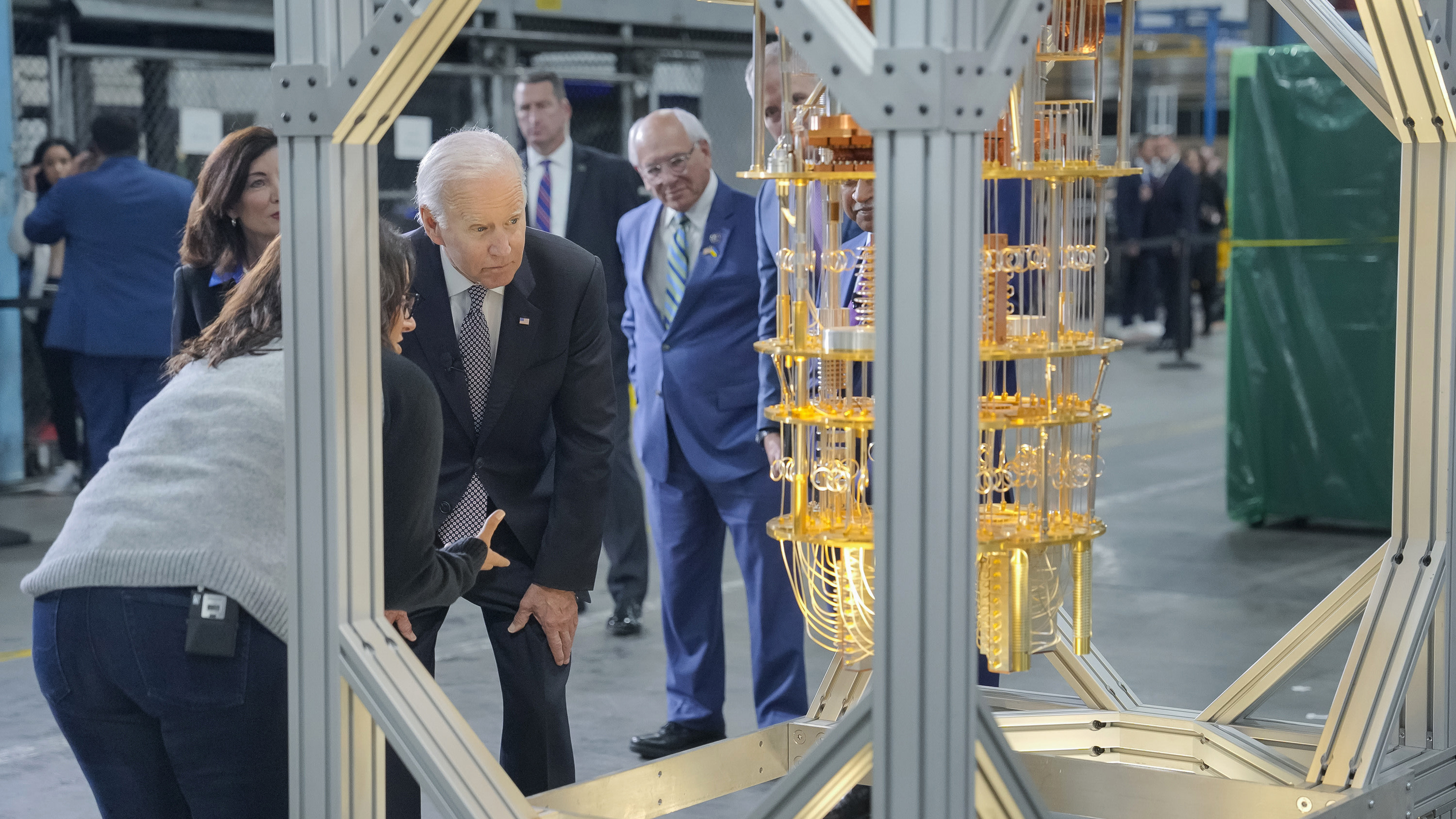 Joe Biden in an industrial-looking facility, with several other individuals looking on and visible in the background, bends to observe a well-lit, intricate gold quantum computer hanging in a metal frame.